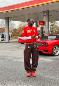 Červené tričko s potla�čou Ferrari a bielymi rukávmi, hnedé široké nohavice a červené topánky. Doplnky zahŕňajú retiazky a slnečné okuliare. V pozadí čerpacia stanica.