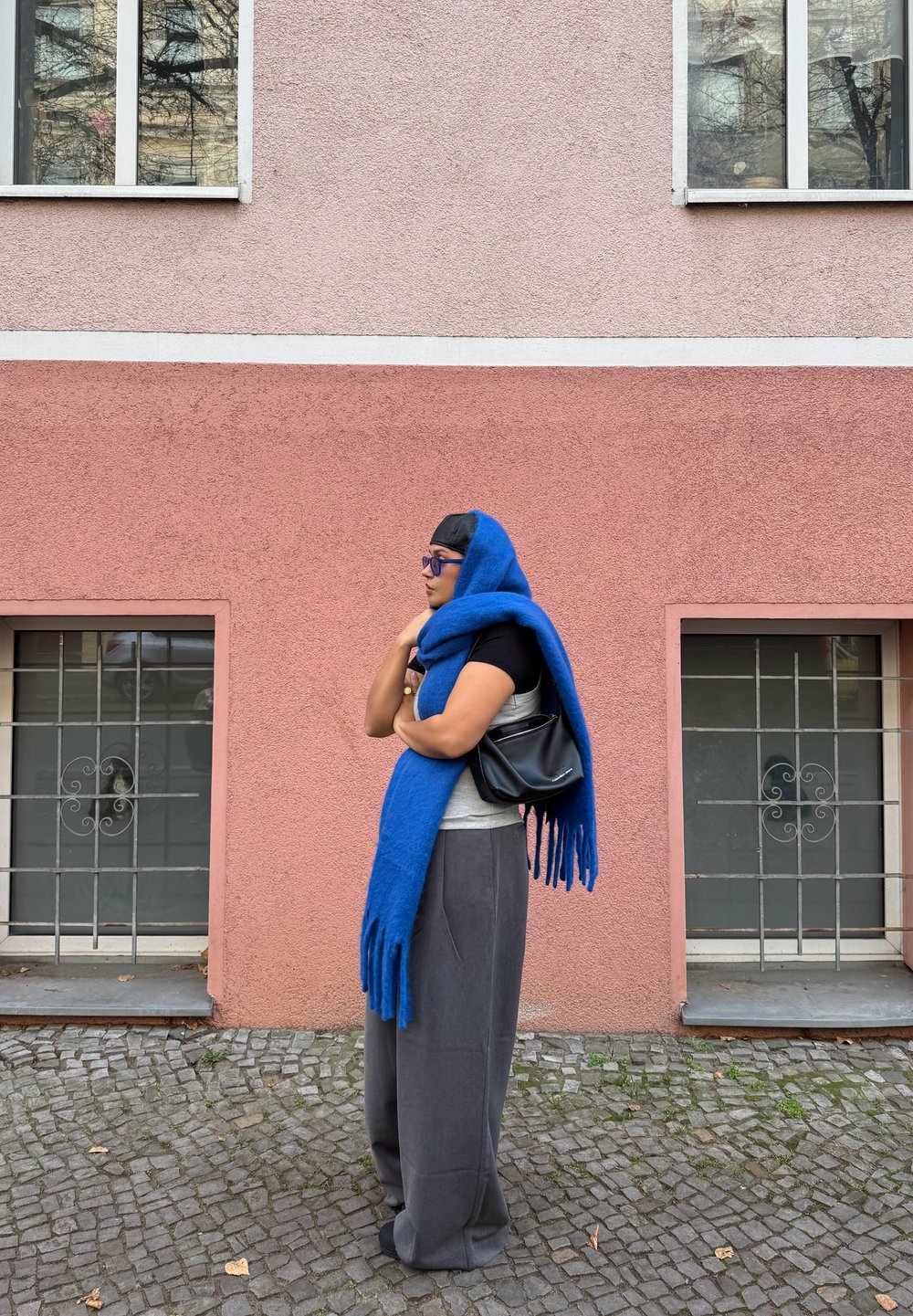 Écharpe en laine bleue avec franges, pantalon gris à jambes larges, haut gris et sac à main noir, devant un mur rose avec des fenêtres et un pavé en pierre.