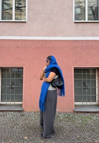Écharpe en laine bleue avec franges, pantalon gris à jambes larges, haut gris et sac à main noir, devant un mur rose avec des fenêtres et un pavé en pierre.