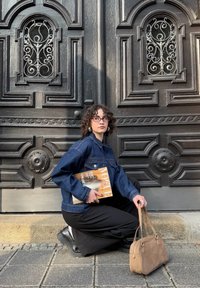 Tumši zila džinsu jaka, melnas bikses, tur rokā žurnāls. Brūna zamšāda rokassoma ar apaļām detaļām. Fons ar greznām melnām durvīm.