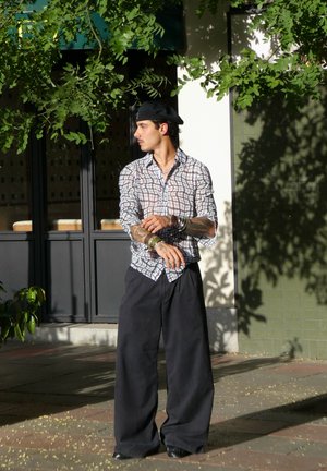 Camisa de cuadros clara, pantalones negros amplios, múltiples pulseras de plata y una boina negra. De pie sobre una acera con vegetación cercana.