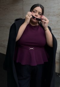 Une femme porte un haut peplum violet avec une boucle de ceinture argentée, un pantalon noir et un manteau noir drapé sur son épaule, tenant des lunettes de soleil.