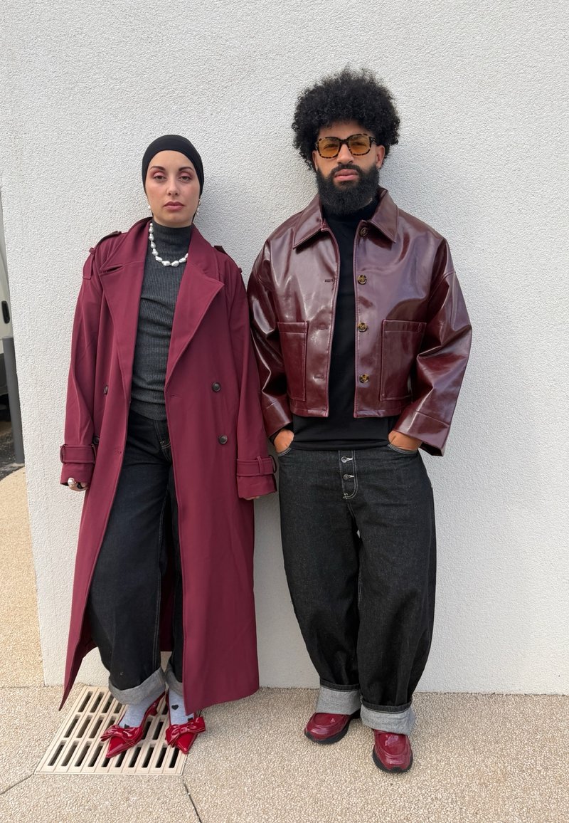 Deux adultes se tiennent contre un mur blanc, vêtus de tenues noires, bordeaux et grises ; l'un porte un long manteau et l'autre une veste en cuir avec des lunettes de soleil.