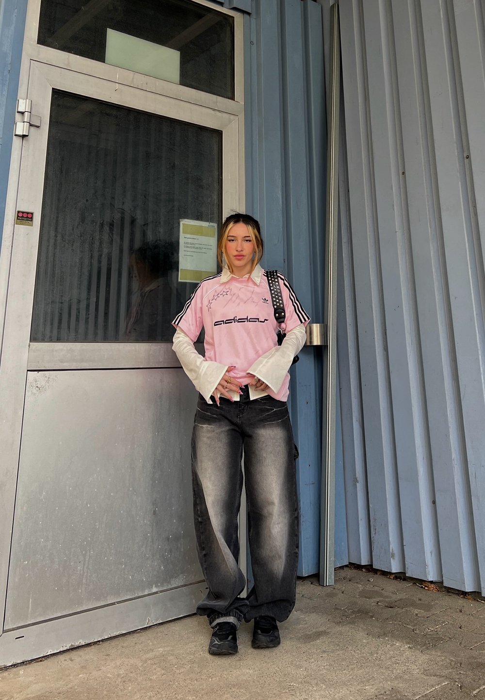 Model nosi roza Adidas majico s črnimi črtami, črne prevelike kavbojke in črne superge, stoji ob kovinskih vratih z modrim panelom.