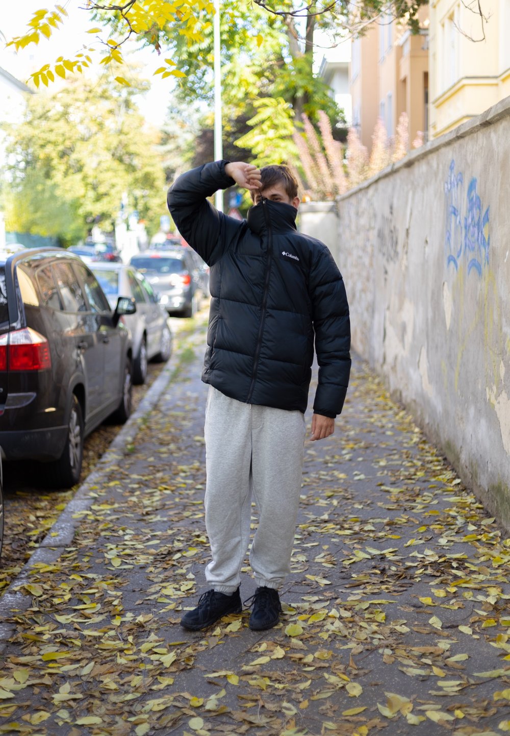 Casaco puffer preto com colarinho alto, calças de moletom cinzas e sapatilhas pretas, de pé numa calçada coberta de folhas.