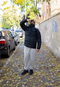 Casaco puffer preto com colarinho alto, calças de moletom cinzas e sapatilhas pretas, de pé numa calçada coberta de folhas.