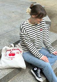 Ριγέ μακρυμάνικο πουκάμισο σε μαύρο και λευκό, με κόκκινες κεντητές λεπτομέρειες, συνδυασμένο με ανοιχτό μπλε τζιν και μαύρα παπούτσια Converse. Καμβάς τσάντα με γραφικό κερασιού και κείμενο.