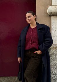 En modell i en marinblå kappa över en vinröd mjuk tröja, ihop med bruna rutiga byxor. Håller en svart väska, står mot en bordeauxfärgad vägg.