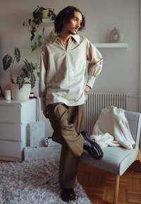Lichtbeige oversized shirt met een puntige kraag en knoopmanchetten, gecombineerd met olijfgroene broek. De schoenen zijn zwart en de achtergrond bevat planten en meubels.