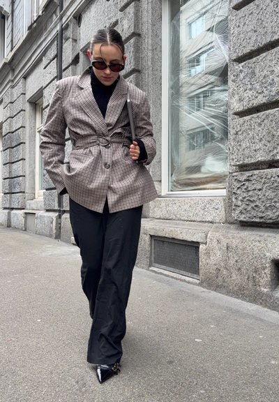 Mujer con blazer a cuadros con cinturón, pantalones negros y zapatos puntiagudos caminando por la acera de la ciudad junto a un edificio de piedra con una ventana grande.