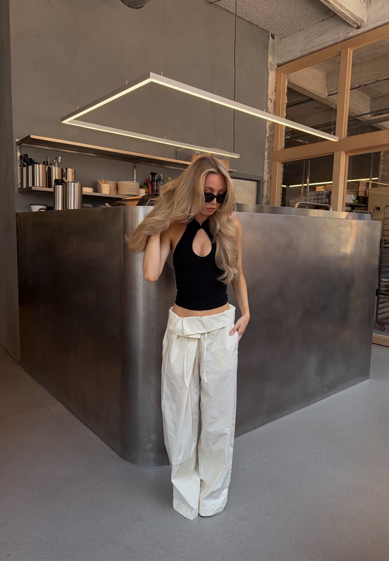 Femme aux longs cheveux blonds portant un haut noir sans manches et un pantalon blanc large, debout à l'intérieur près d'un comptoir de cuisine moderne en métal.