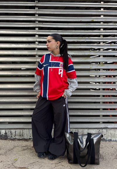 Joven con coleta lleva una camiseta de fútbol roja de Noruega, sudadera gris con capucha, pantalones negros Adidas, está de pie junto a una pared de metal con una bolsa negra en el suelo.