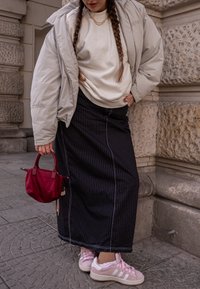 Casaco oversized acolchoado cinza, sweatshirt branca, saia maxi preta riscada, ténis cor-de-rosa, mala vermelha, fundo de pedra texturizada.