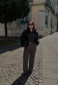 Zwarte leren jas, grijze trui, wijde beige broek en een zonnebril. De achtergrond toont een kasseien straat en een gebouw met balkons.