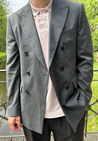 Blazer grigio doppiopetto con bottoni neri, rever a contrasto e una tasca sul petto, indossato sopra una maglietta polo rosa chiaro.