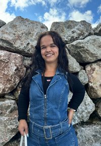 Σαλοπέτα από τζιν με μπροστινό φερμουάρ, χωρίς μανίκια, και ζώνη στη μέση. Μαύρη μπλούζα με μακριά μανίκια από κάτω. Φυσικό υπόβαθρο από πέτρα.