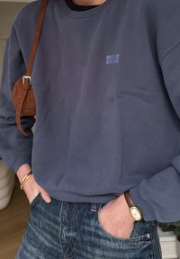 Personne portant un sweat-shirt marine avec le logo "American Vintage", un jean bleu, une montre marron, des bracelets dorés et un sac à bandoulière en daim marron.