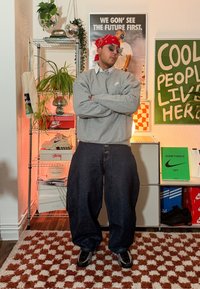 Sweat-shirt gris avec un petit logo, jean bleu oversize et baskets noires. Accessoires comprend des lunettes de soleil et un bandana rouge. En arrière-plan, étagères et affiches.