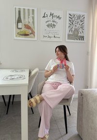 Donna seduta su una sedia grigia chiara, vestita con una camicia bianca e pantaloni a scacchi rosa, tiene in mano una tazza a righe. Vicino, un tavolo bianco con dei sottopiatti decorati.