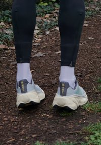 Ténis de desporto cinzentos com solas texturizadas, com "MORE" em amarelo na parte de trás, utilizados com meias brancas e calças justas pretas num caminho de terra.