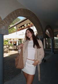 Beige trenčkot přes bílý top a texturovanou bílou sukni. Modelka drží hnědou semišovou tote tašku. V pozadí kamenné oblouky a venkovní sezení.