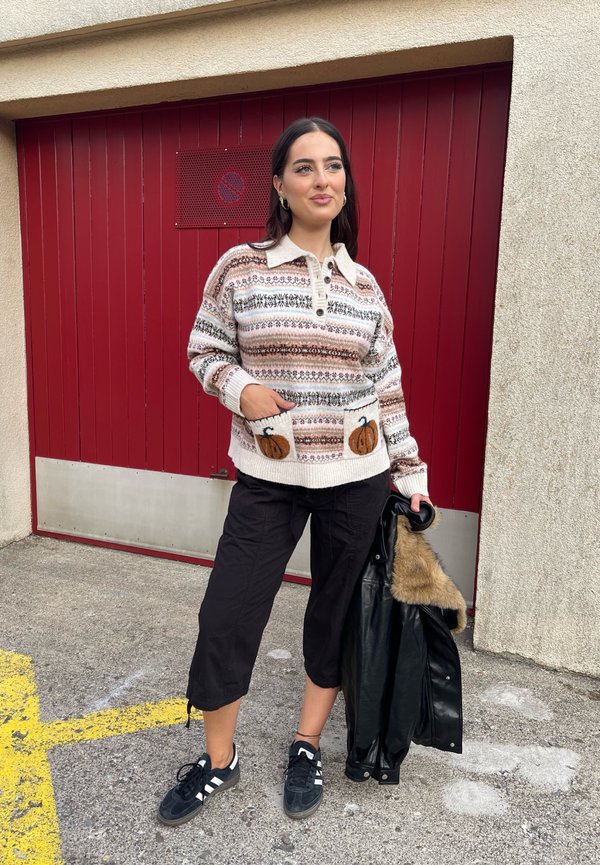 Mujer de pie al aire libre vistiendo un jersey estampado con diseños de calabazas, pantalones negros recortados y sosteniendo una chaqueta negra con cuello de piel.