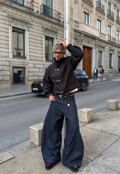 Persona con chaqueta negra, pantalones oscuros y anchos, sombrero peludo y anillos, de pie en una acera de la ciudad cerca de una farola y edificios.