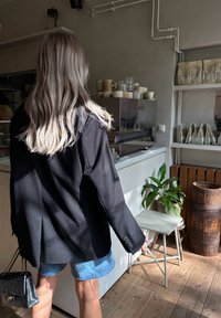 Čierny blejzer s voľným strihom, skombinovaný s poškodenými džínsovými šortkami. Doplnky zahŕňajú prešívanú čiernu kabelku. Viditeľná drevená podlaha.