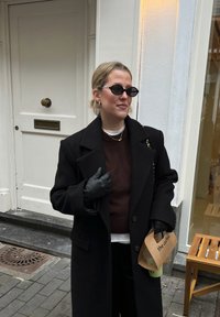 Femme portant un manteau noir, des lunettes de soleil et des gants, tenant un café à emporter, debout dans une rue près d'une porte blanche et d'un banc en bois.