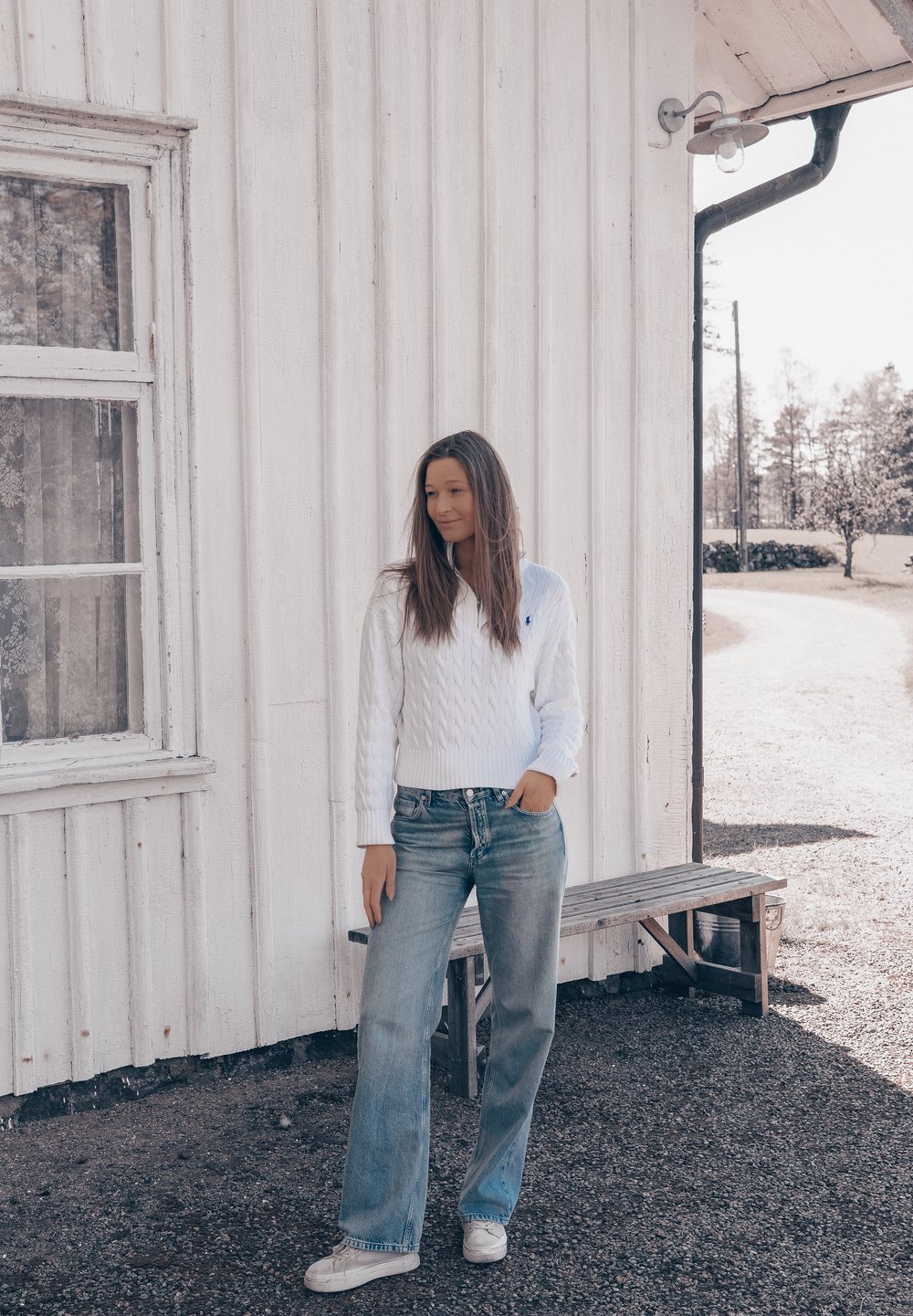 Maglione bianco a trecce con scollo a V, abbinato a jeans azzurri a vestibilità rilassata e scarpe bianche casual, in piedi contro un muro di legno.
