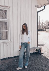 Maglione bianco a trecce con scollo a V, abbinato a jeans azzurri a vestibilità rilassata e scarpe bianche casual, in piedi contro un muro di legno.