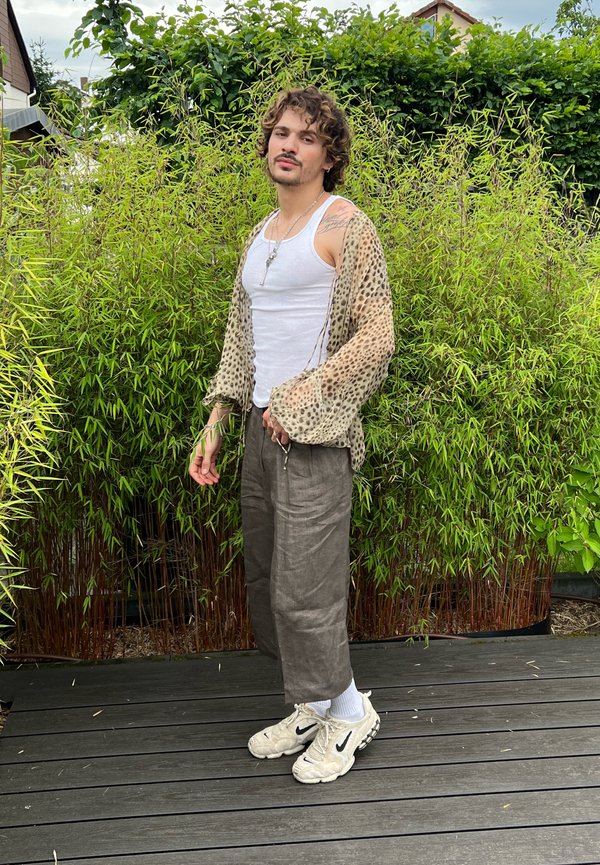 Tânăr cu păr creț, purtând un maiou alb, cămașă transparentă cu imprimeu leopard, pantaloni largi și adidași albi, stând pe o terasă din lemn, cu plante verzi pe fundal.