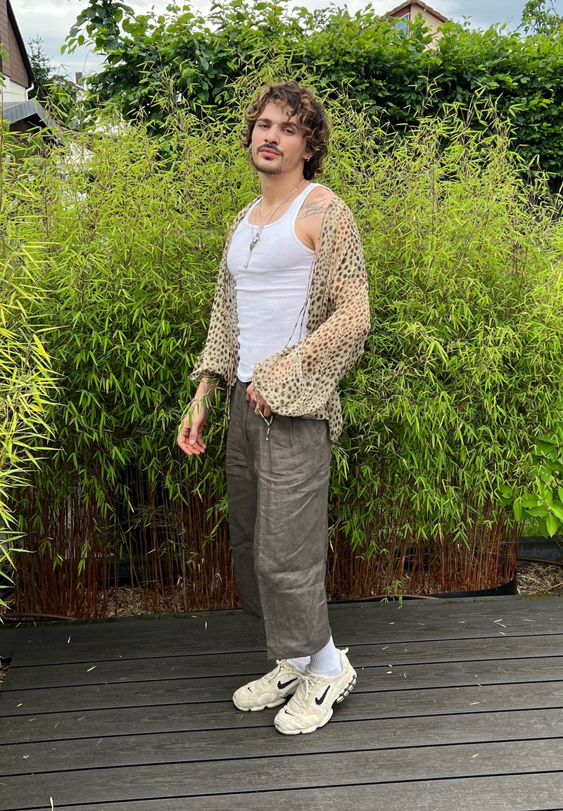 Jeune homme aux cheveux bouclés portant un débardeur blanc, une chemise transparente à imprimé léopard, un pantalon ample et des baskets blanches, debout sur un pont en bois avec un fond de plantes vertes.
