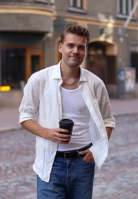 Bílá lněná košile přes bílý tílko, modré džínové kalhoty, držící černý hrnek s kávou, stojící na dlažební uličce.
