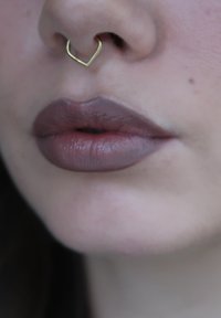 Detalle de labios con lápiz labial púrpura apagado y un aro septum en forma de corazón dorado sobre piel clara.