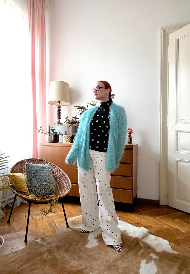 Γυναίκα με κόκκινα μαλλιά και γυαλιά στέκεται σε ένα δωμάτιο φορώντας ένα μπλε χνουδωτό ζακέτα, μαύρο τοπ με πουά, άσπρο παντελόνι με πουά και ριγέ παπούτσια.