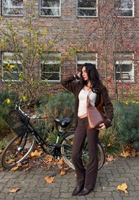 Brown leather shoulder bag with a geometric shape, worn with a white top and dark brown trousers, beside a black bicycle and foliage.