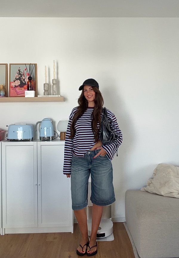 Mujer que lleva una camiseta de manga larga a rayas, shorts vaqueros, gorra negra y sandalias, de pie en una sala de estar con armarios blancos y un sofá beige.