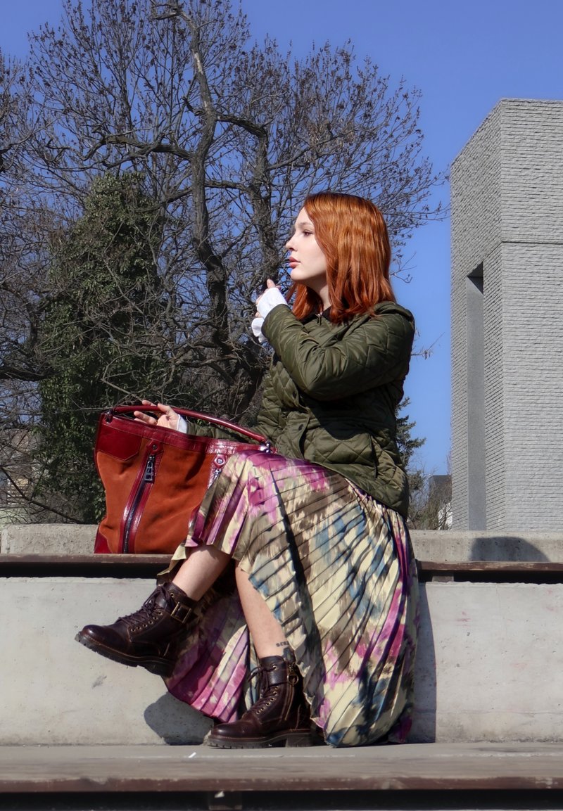 Donna con capelli rossi, giacca verde e gonna tie-dye seduta su una panchina di cemento, che tiene una grande borsa marrone, guardando di lato all'aperto.