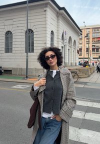 Modell bär en ljusgrå rutig kappa över en mörk väst, i kombination med en vit skjorta och blå jeans. Solglasögon fulländar looken.