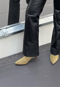 Zwarte leren flarebroek gecombineerd met gele suède pumps met een dunne houten hak. De achtergrond weerspiegelt een metallic oppervlak.