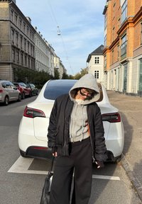Inimesed hallis kapuutsiga ja mustas jope koos prillidega seisab valge pargitud auto ees linnatänaval, kus on telliskivist hooned.