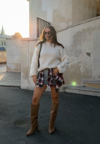 Mujer con gafas de sol, suéter blanco, camisa de cuadros atada a la cintura, pantalones cortos de mezclilla y botas marrones hasta la rodilla, parada cerca de unas escaleras de piedra al aire libre.