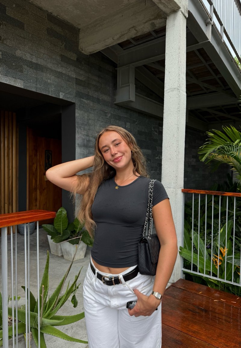 Camiseta negra ajustada, pantalones blancos de talle alto y un bolso negro con asa de cadena. Rodeada de vegetación y arquitectura moderna.