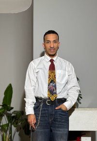 Homme portant une chemise blanche, une cravate colorée à motifs, un jean foncé avec une chaîne, debout à l'intérieur la main dans la poche près des plantes et d'une table.