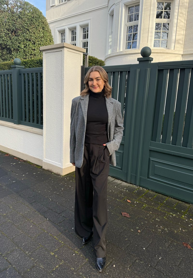 Grå blazer over en sort rullekrave, kombineret med sorte bukser med vide ben og spidse sorte sko. Belagt gangsti foran en grøn port.