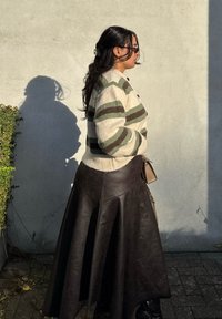 Pletená kardigán s zelenými a hnedými pruhmi, zapínaním na gombíky a rebrovaným lemom, skombinovaná s objemnou hnedou koženou sukňou a čiernymi čižmami.