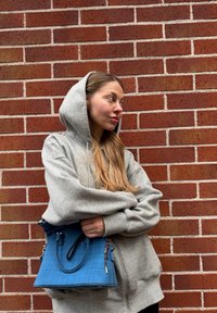 Jeune femme en sweat à capuche gris tenant un sac à main bleu, debout contre un mur de briques rouges, regardant sur le côté avec les bras croisés.