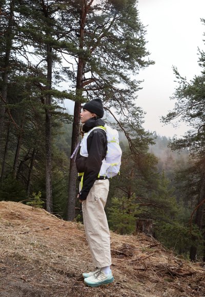 Persona con una chaqueta negra, pantalones beige y un gorro negro, con una mochila blanca con detalles en amarillo, de pie en una zona boscosa.