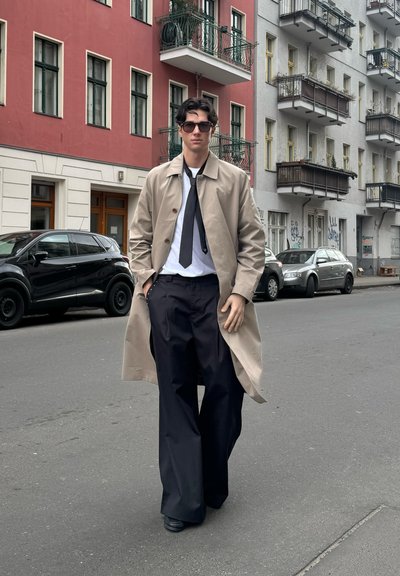 Abrigo beige sobre una camisa blanca y corbata negra, combinado con pantalones anchos negros. Accesorios incluyen gafas de sol oscuras. Entorno urbano.